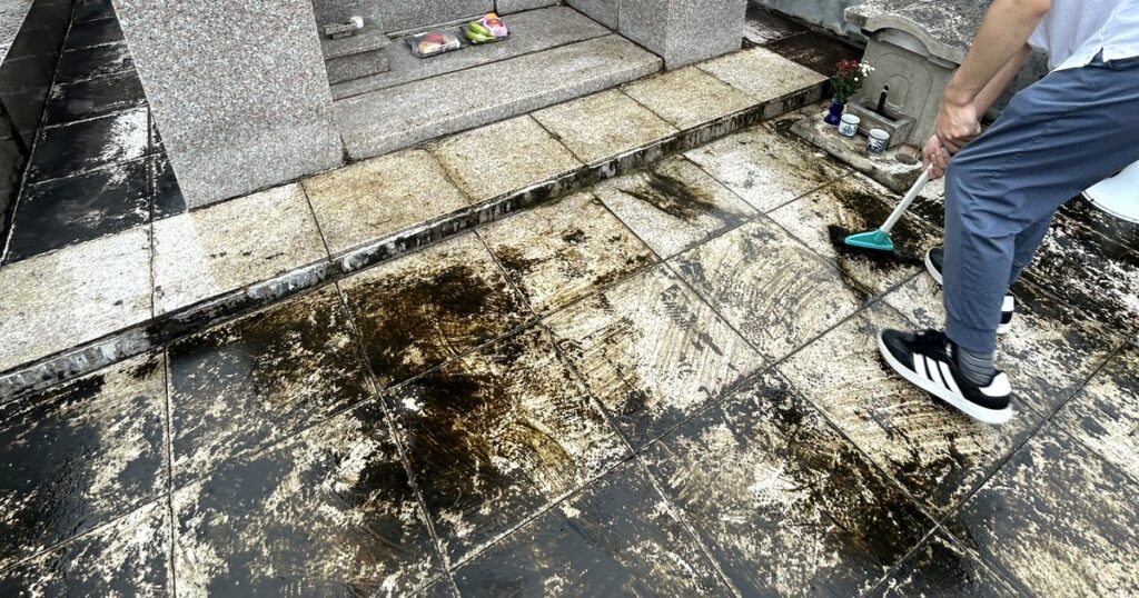 お墓の床のタイルに黒いコケが広がり、ほうきで掃いている様子